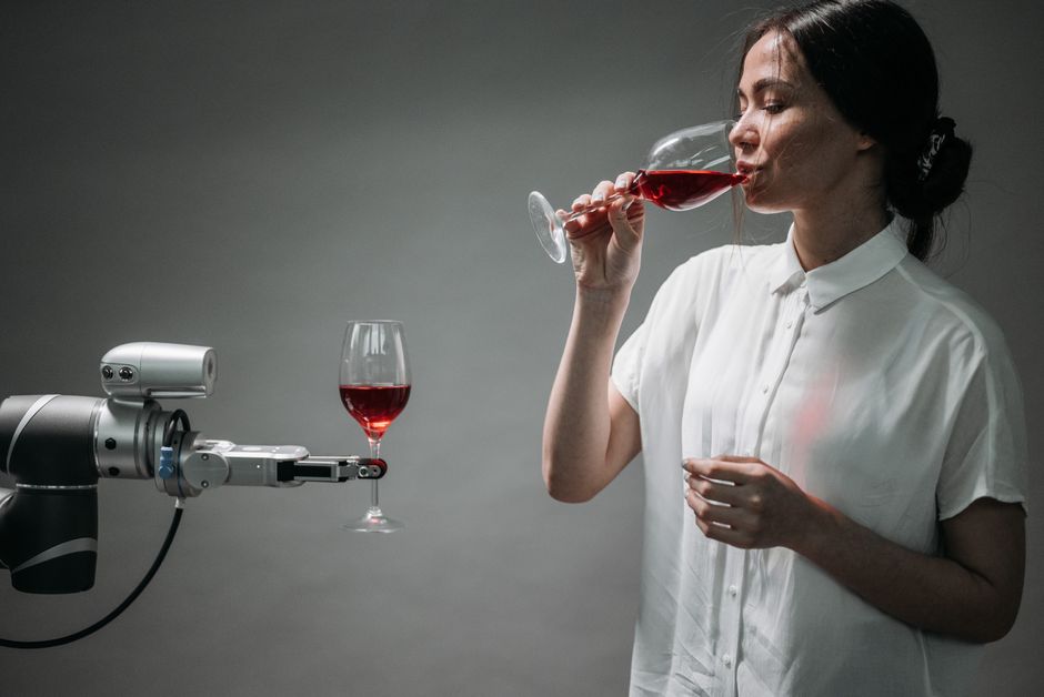 Una mujer con una camisa blanca bebiendo vino tinto interactúa con un brazo robótico que sostiene una copa de vino.