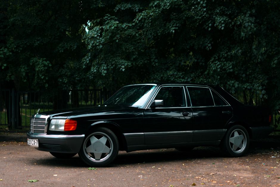 Un coche vintage Mercedes-Benz W126 negro aparcado en un entorno exterior sombreado.