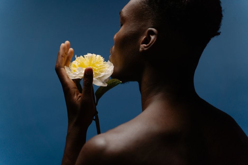 Hombre sin camisa sostiene una flor amarilla sobre un fondo azul profundo, destacando el contraste.