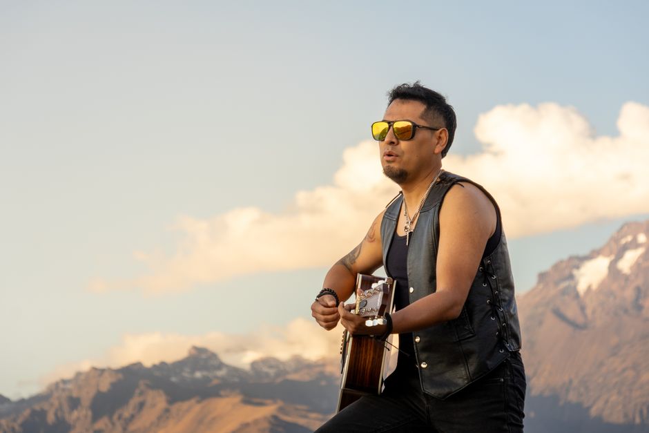 Un músico solista, con gafas de sol, toca la guitarra con un impresionante paisaje montañoso de fondo.