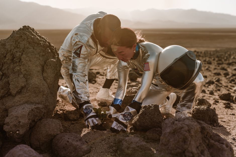Dos astronautas plantando una plántula en un terreno desértico rocoso, simbolizando la colonización espacial.