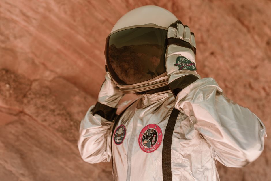 Close-up de un astronauta vistiendo un traje espacial plateado en un entorno rocoso al aire libre.