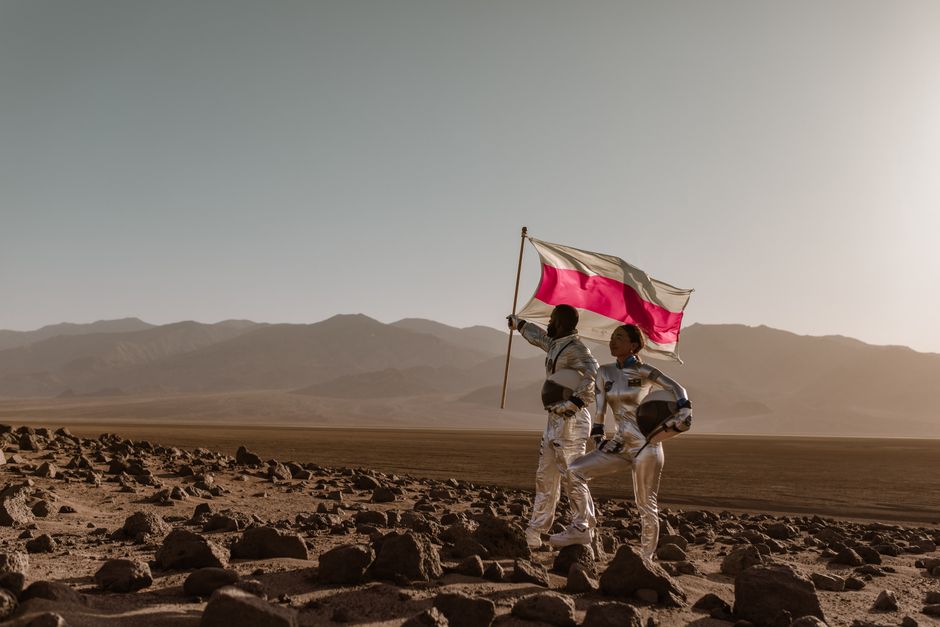 Dos astronautas con trajes están de pie en un terreno rocoso, sosteniendo una bandera contra un fondo montañoso.