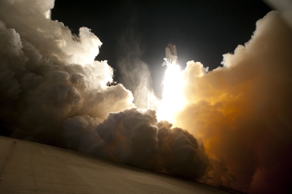 Una imagen dramática de un transbordador espacial despegando hacia el cielo nocturno, rodeado de humo denso y luz intensa.