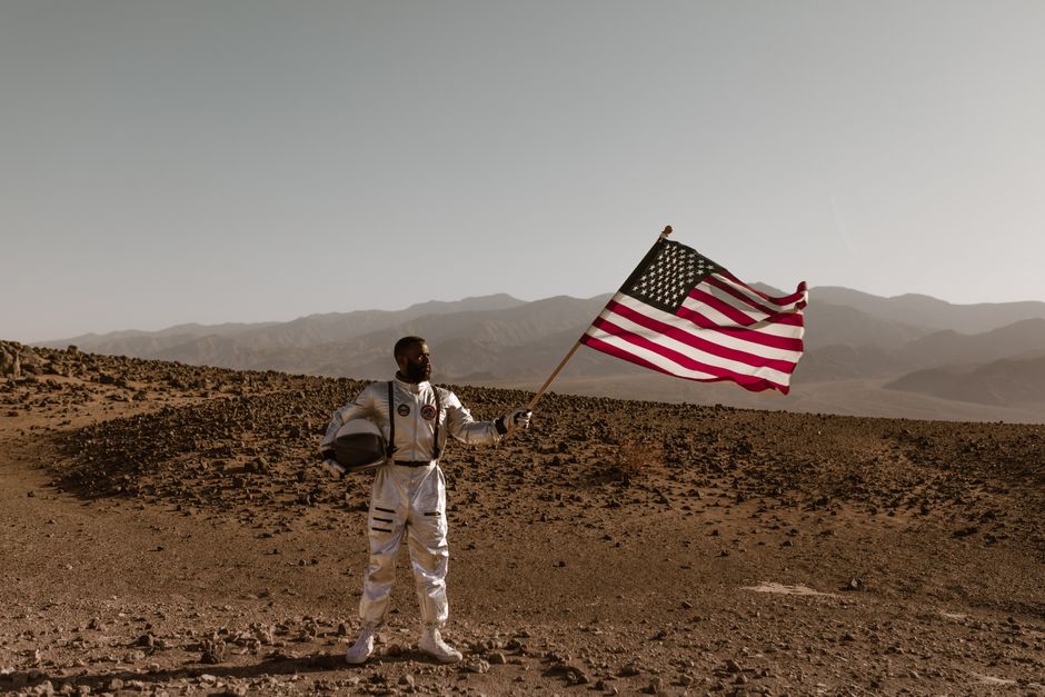 Un astronauta en un traje espacial se encuentra en un paisaje desértico sosteniendo una bandera americana.