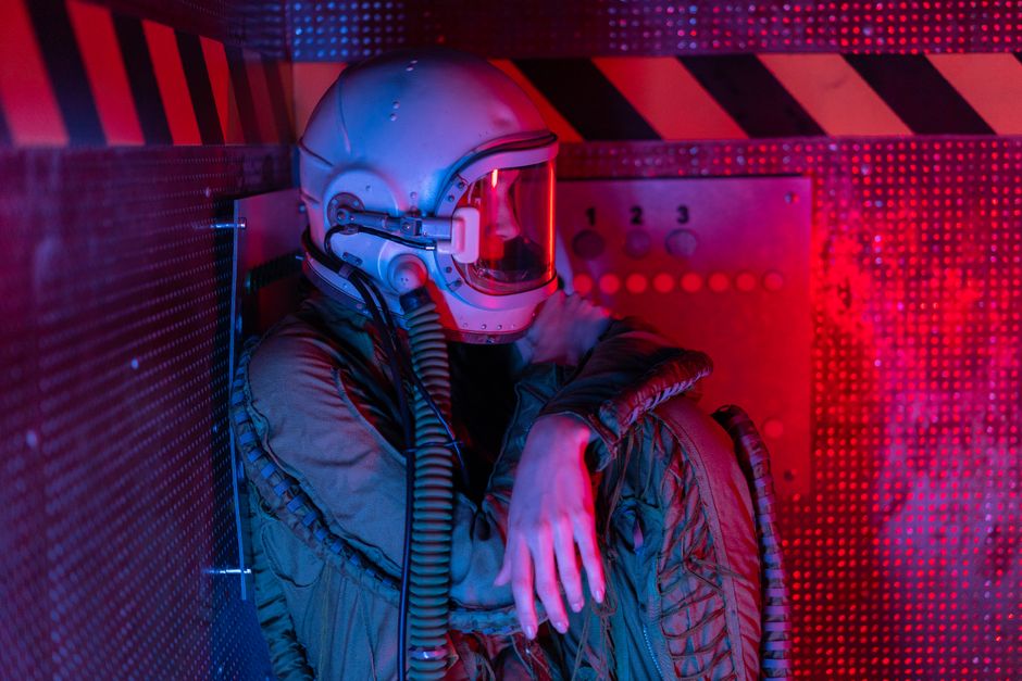 Una astronauta pensativa en escafandra se sienta en un espacio futurista con poca luz, rodeada de luces rojas y moradas.