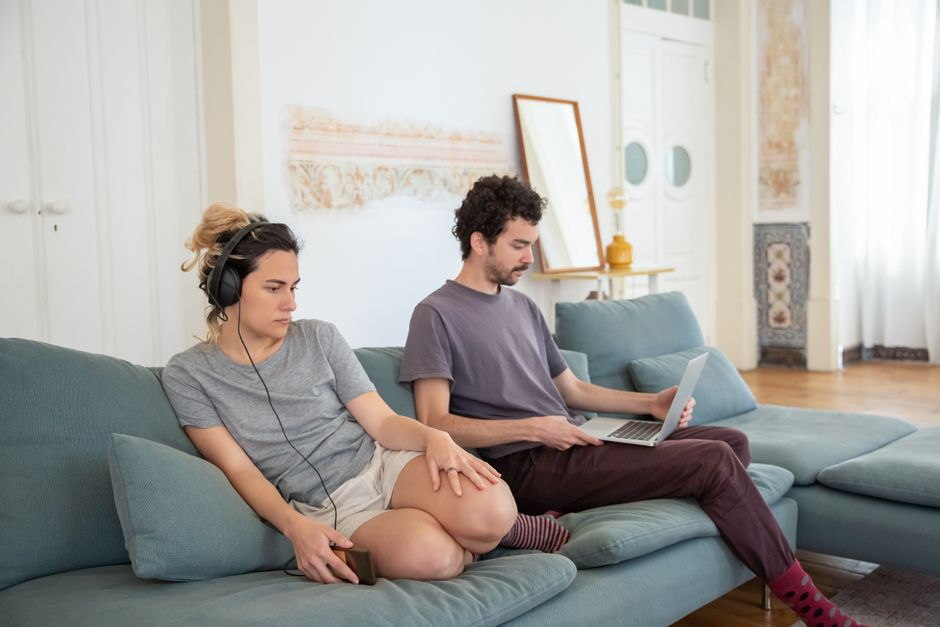 Una pareja relajándose en un sofá con auriculares y laptop, creando un ambiente acogedor en casa.