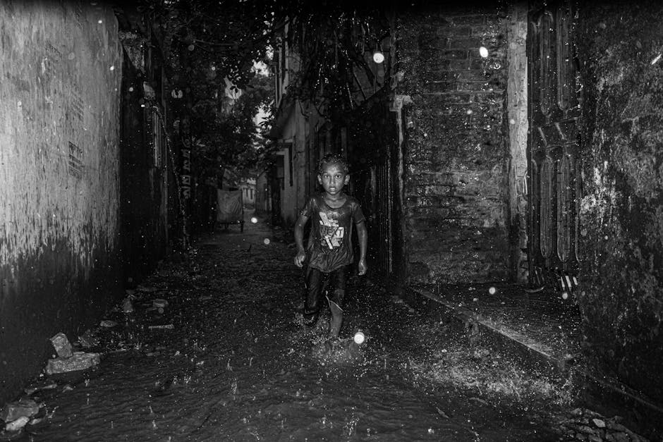 Un niño pequeño corre alegremente por un callejón urbano oscuro y lluvioso, capturado en una dramática imagen en blanco y negro.