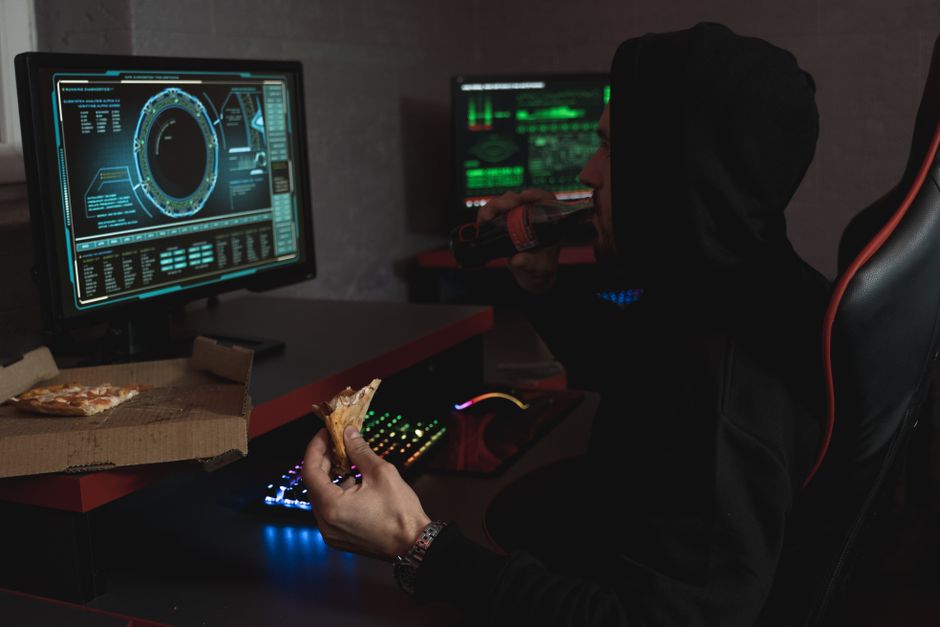 Hacker con sudadera negra comiendo pizza y bebiendo refresco mientras analiza datos en múltiples pantallas de ordenador.