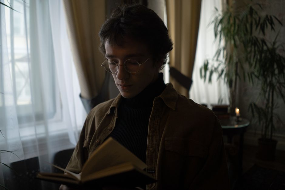 Un joven con gafas absorto en la lectura de un libro junto a la ventana con luz suave.
