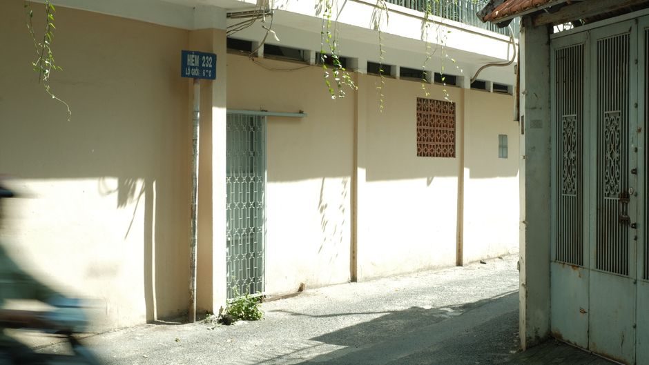 Callejón iluminado por el sol en Vietnam con sombras y un cartel que dice 'Hẻm 232'.