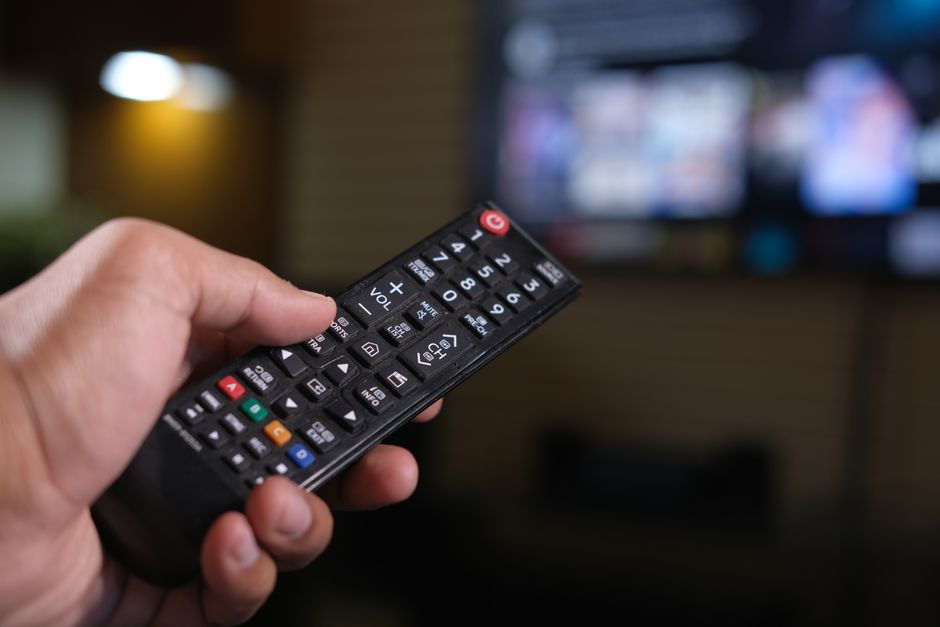 Persona sosteniendo un control remoto con una pantalla de TV desenfocada al fondo.