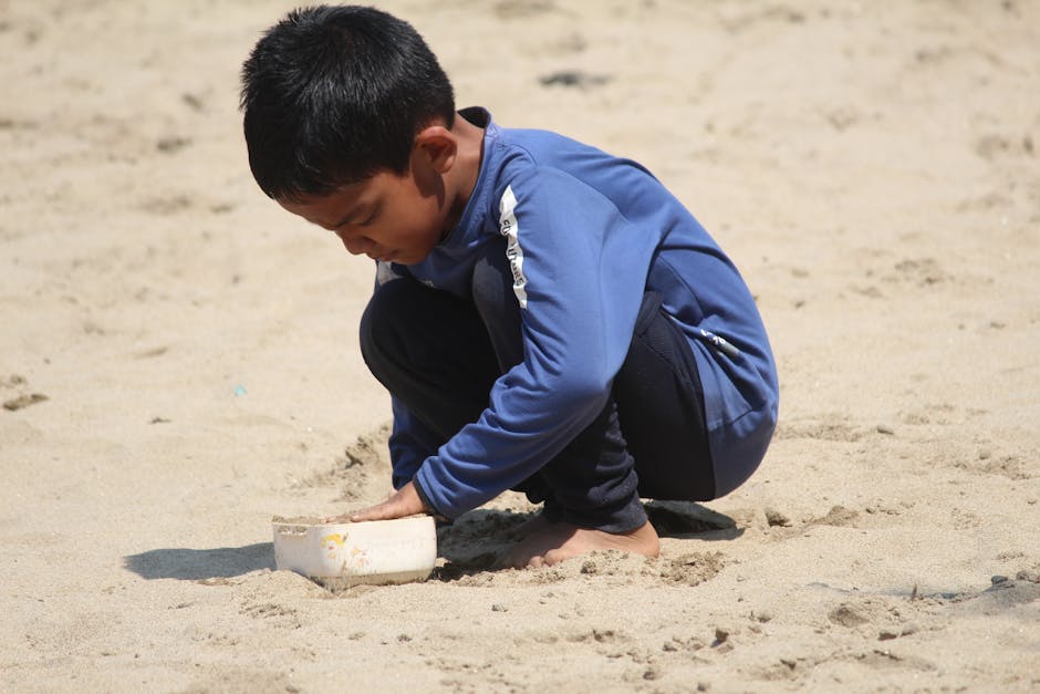 Un niño con una chaqueta azul se concentra en jugar con arena en la playa en un día soleado.