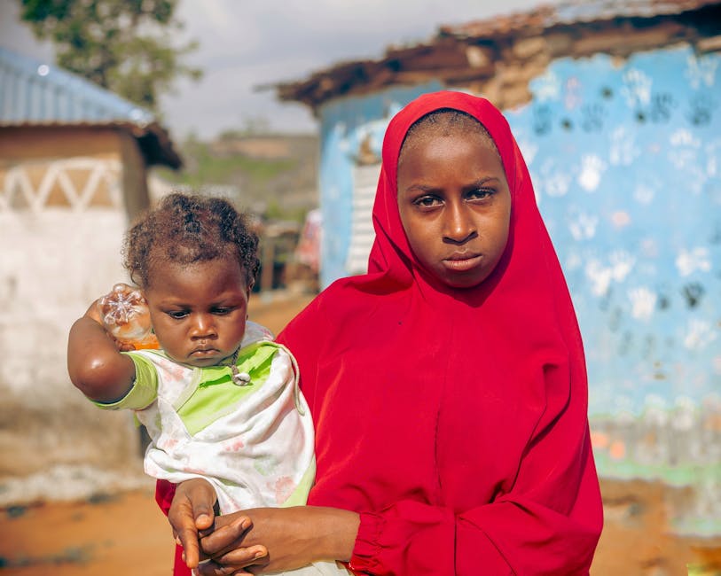 Una mujer con un hijab rojo sostiene a un niño pequeño frente a un vibrante paisaje rural.