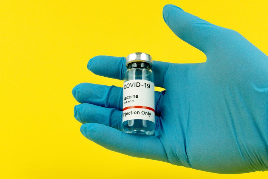 Una mano enguantada sostiene un vial de vacuna COVID-19 frente a un fondo amarillo brillante.