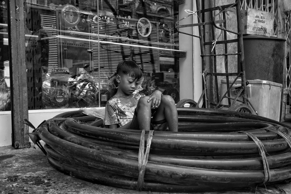 Fotografía en blanco y negro de un niño sentado en unas tuberías enrolladas frente a una tienda, que muestra una escena de la vida urbana.
