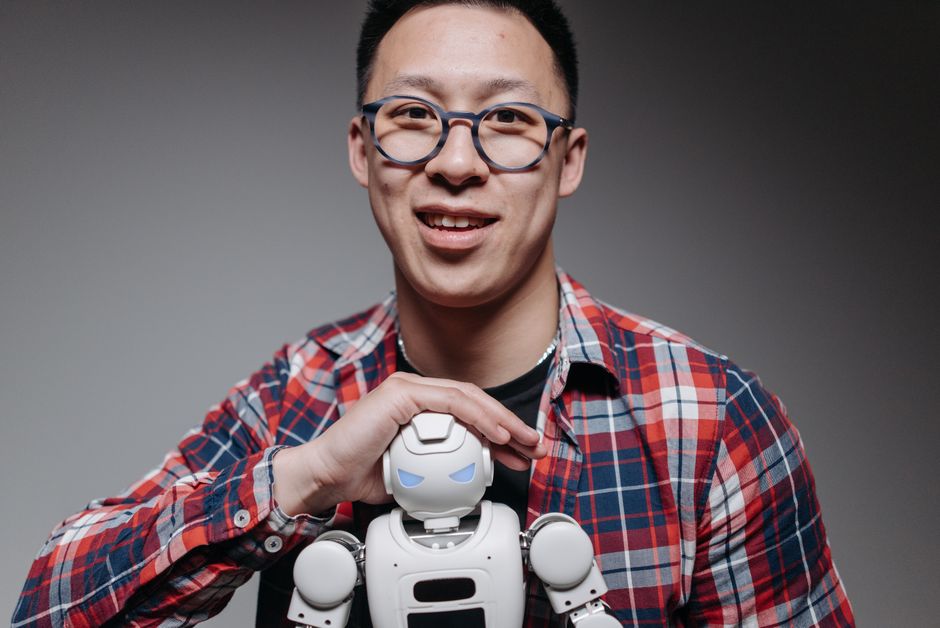 Hombre asiático sonriendo, sosteniendo un pequeño robot de juguete durante una sesión de estudio. En interiores, con gafas y camisa de cuadros.