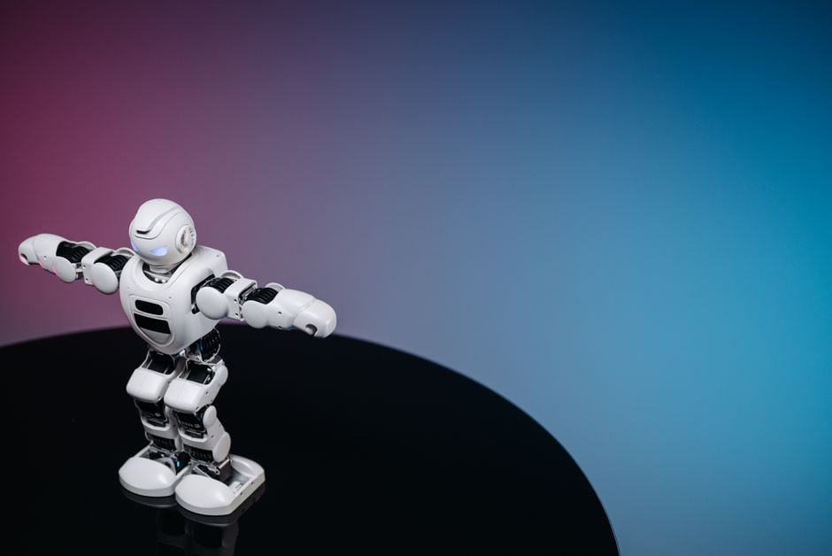 Un robot humanoide blanco de pie sobre una superficie negra reflectante en un entorno de estudio con un fondo de degradado azul y rosa.