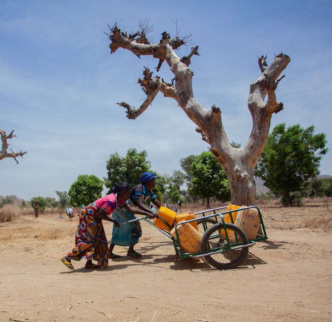 Dos mujeres empujan un carro con contenedores de agua en una zona rural seca.