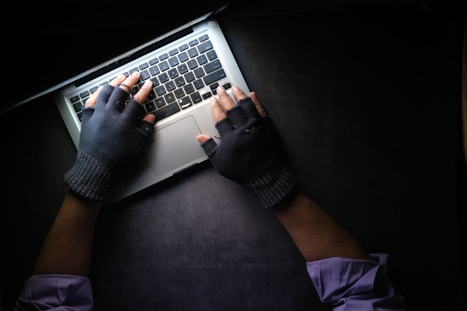 Hacker con guantes sin dedos tecleando en el teclado del laptop desde arriba en un entorno oscuro.