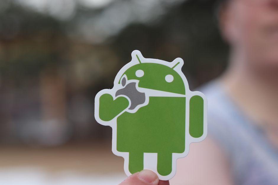 Primer plano de una etiqueta de Android, sosteniendo de manera humorística un logotipo de manzana, con un fondo desenfocado al aire libre.