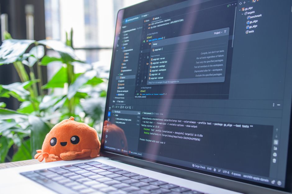 Portátil abierto con editor de código visible, junto a un juguete de peluche naranja y plantas verdes.