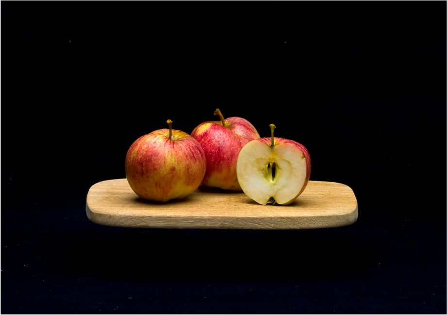 Tres manzanas rojas sobre una tabla de cortar de madera contra un fondo oscuro. Una está cortada por la mitad.