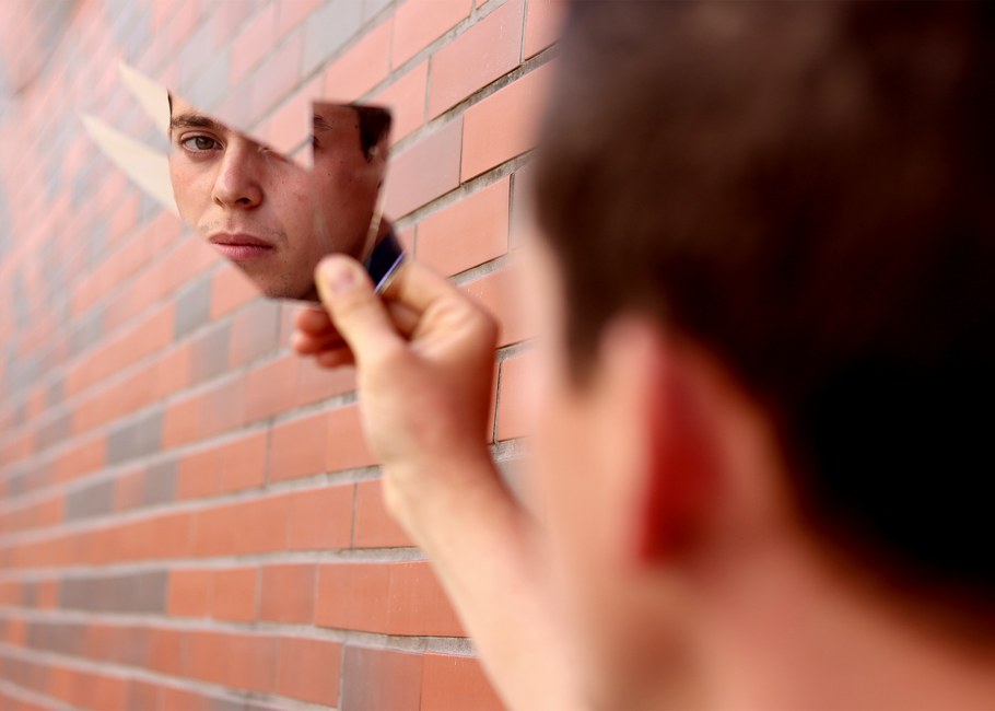 El rostro de un hombre se refleja en un fragmento de espejo sostenido contra una pared de ladrillos.