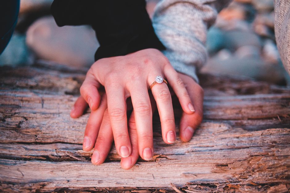 Un primer plano romántico de una pareja tomándose de las manos con un anillo de compromiso sobre un tronco de madera.