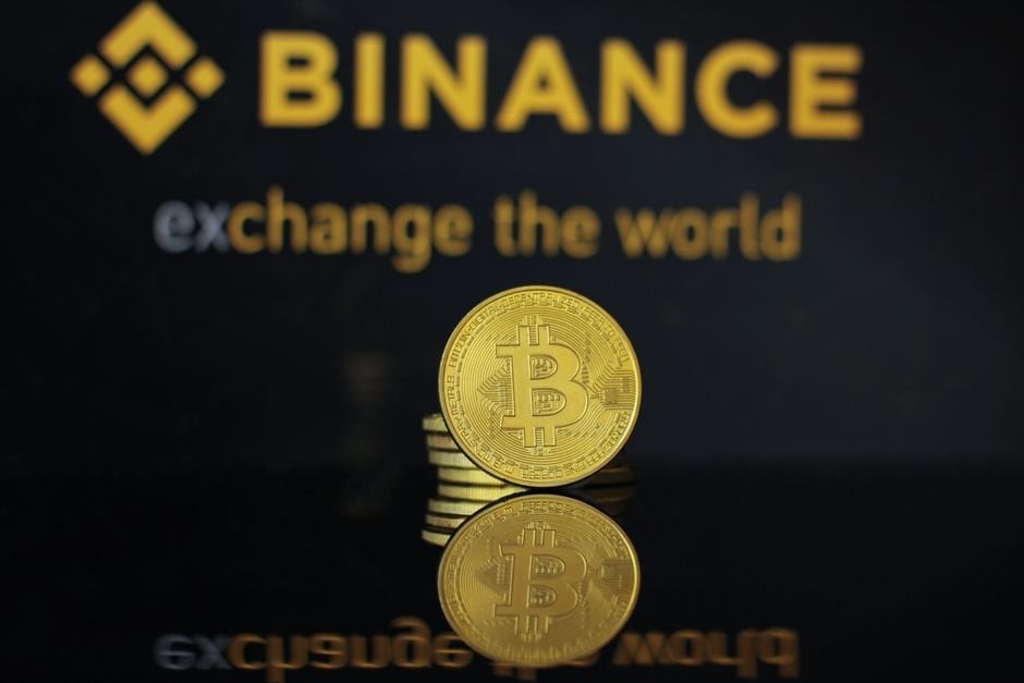 Moneda dorada de Bitcoin sobre monedas apiladas, con el logotipo de Binance reflejado en una superficie oscura.