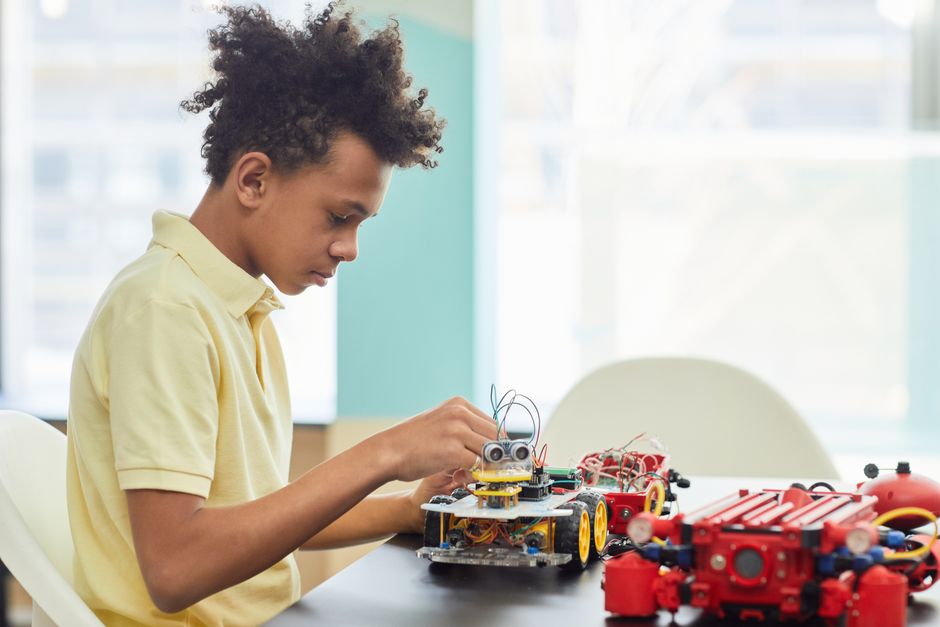 Un niño pequeño concentrado en construir un proyecto robótico en el interior, con coches de juguete y componentes electrónicos.