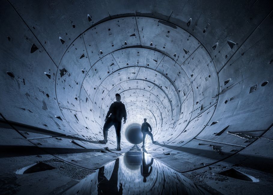 Dos figuras silueteadas exploran un dramático túnel de concreto futurista por la noche.