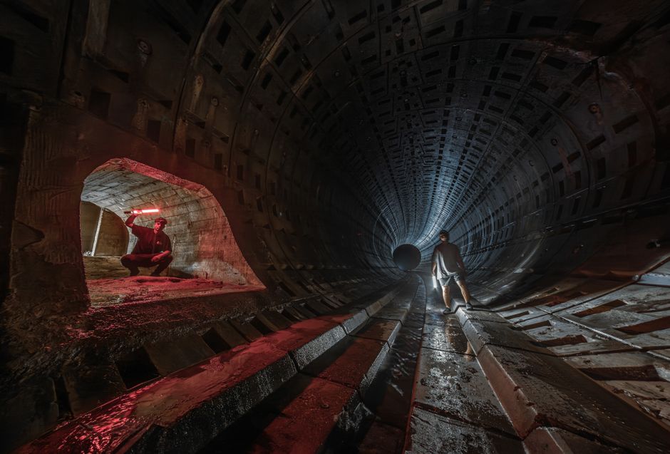 Dos personas exploran un túnel subterráneo tenuemente iluminado con luces rojas, añadiendo una sensación misteriosa y aventurera.