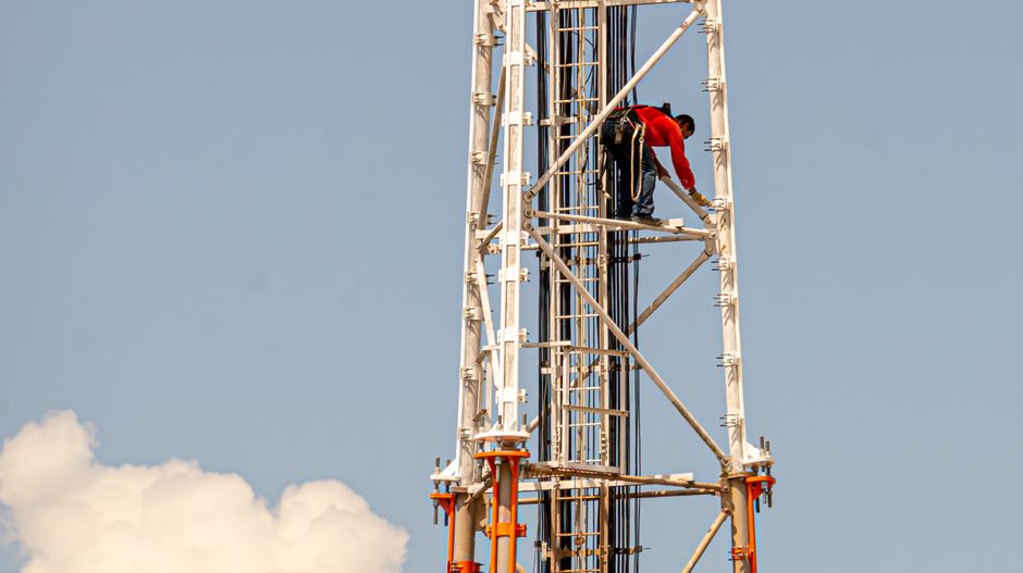 Un trabajador de construcción subiendo una alta torre de comunicación contra un cielo azul claro en Brasil.