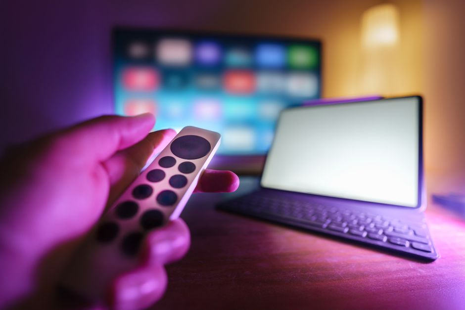 Una mano usando un control remoto para controlar una TV y un portátil en un entorno acogedor, perfecto para relajarse.