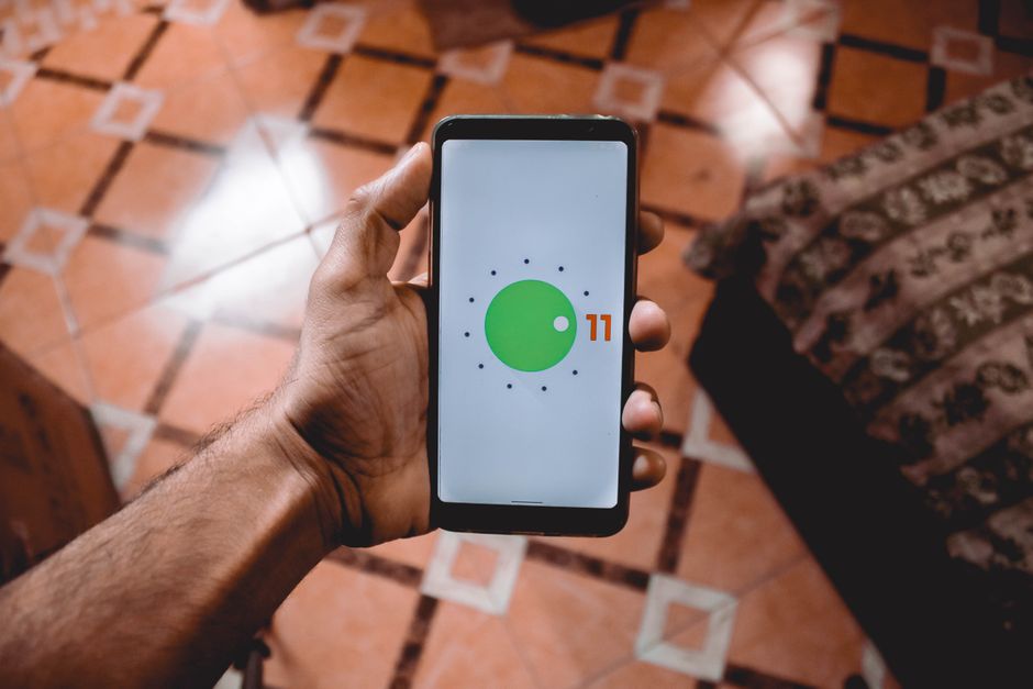Primer plano de una mano sosteniendo un smartphone que muestra la interfaz de Android 11 en el interior sobre un suelo con patrón.
