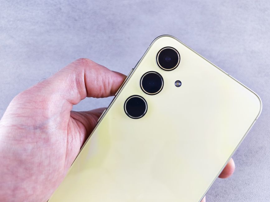 Vista detallada de un smartphone amarillo destacando sus lentes de cámara triple sostenidas por una mano en interiores.