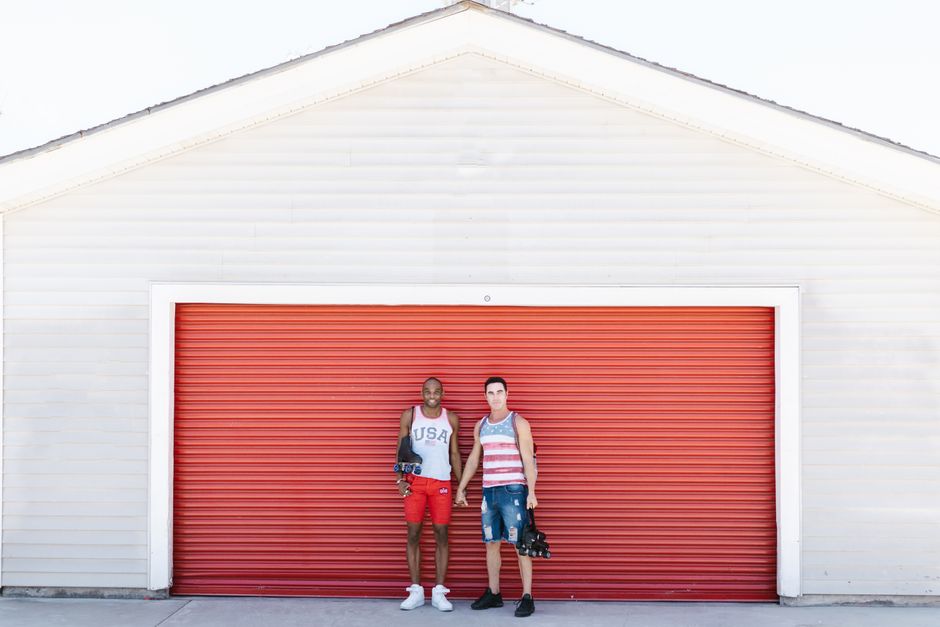 Dos hombres juntos frente a una puerta de garaje roja, luciendo moda de verano.