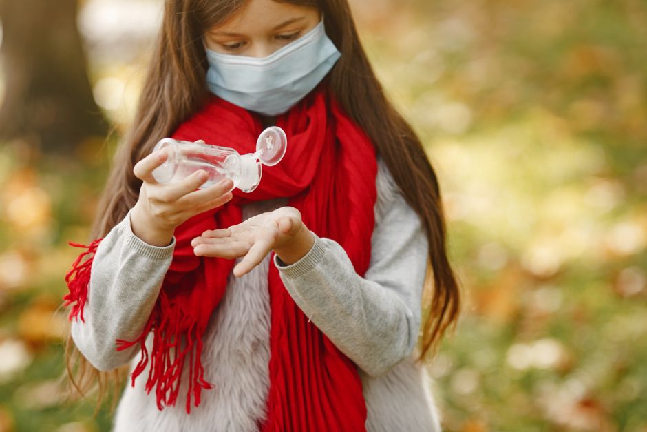 Una niña joven con una máscara y una bufanda roja aplica desinfectante de manos en un parque durante el otoño.
