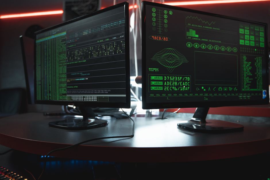 Acercamiento de monitores duales con interfaces de codificación verde en una habitación oscura, destacando temas de ciberseguridad.