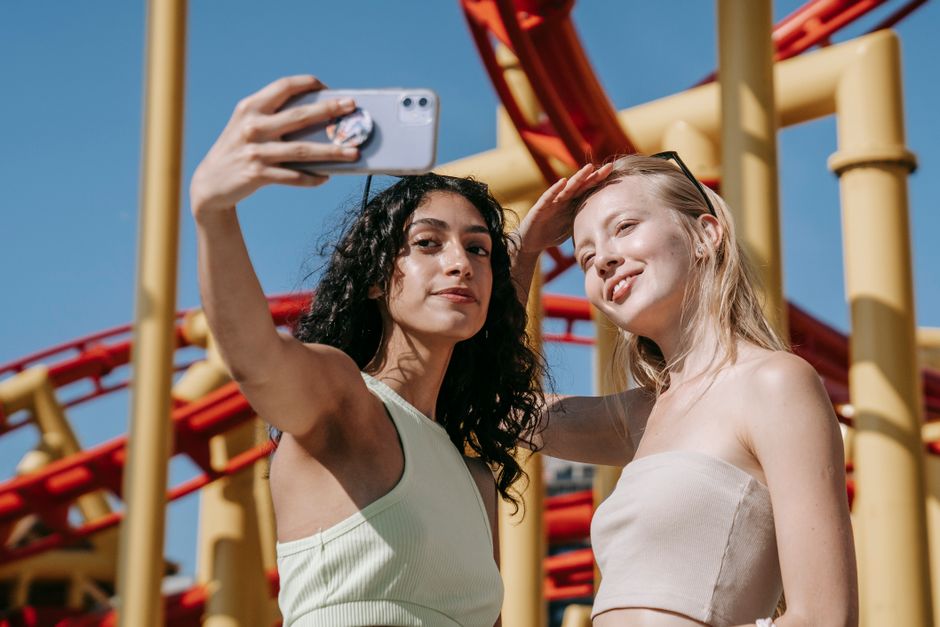 Dos mujeres disfrutando de un día soleado, tomando un selfie en un parque de atracciones con un fondo brillante de montaña rusa.