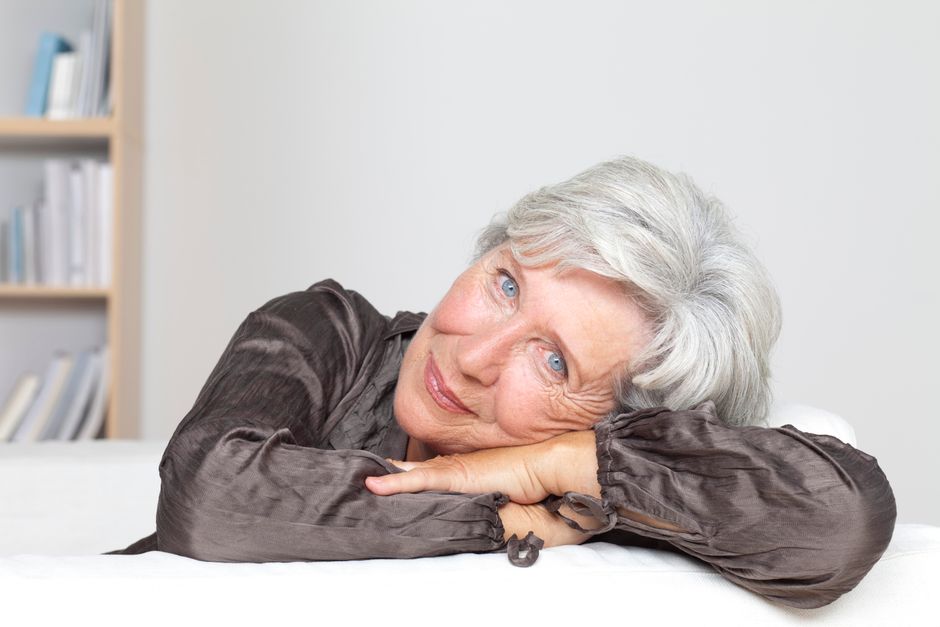 Mujer anciana con cabello canoso sonriendo y apoyada en una mesa en interiores.