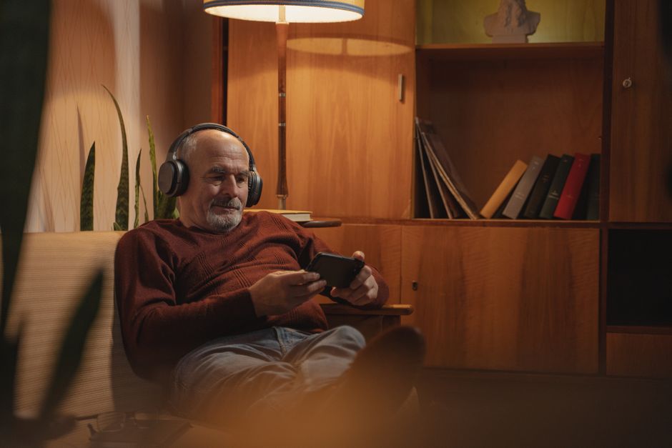 Hombre anciano disfrutando de música con auriculares mientras se relaja con un smartphone en casa.