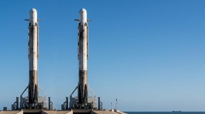 Dos cohetes Falcon 9 de SpaceX idénticos están listos en sus plataformas de lanzamiento sobre un fondo de cielo azul.