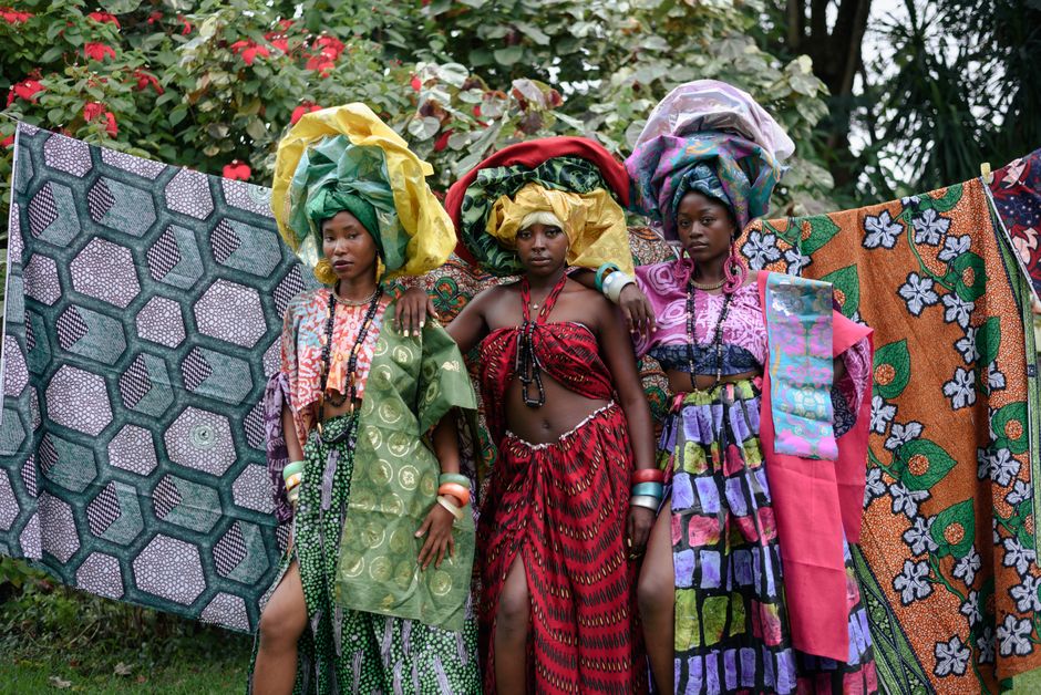 Tres mujeres con coloridos atuendos tradicionales posan al aire libre, mostrando textiles africanos.