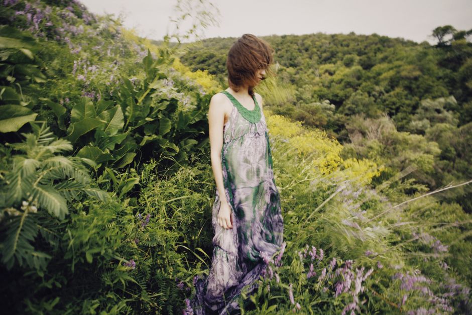 Una mujer serena se encuentra en un prado exuberante y vibrante, vistiendo un vestido fluido, rodeada de plantas silvestres florecientes y naturaleza.