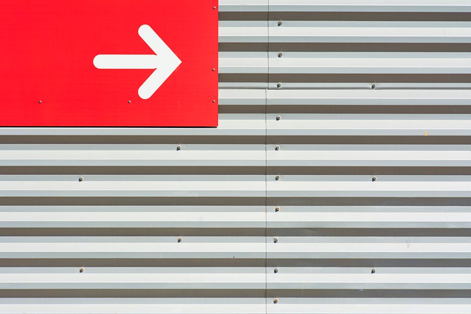 Imagen minimalista que presenta una señal de flecha roja en una pared de metal corrugado.