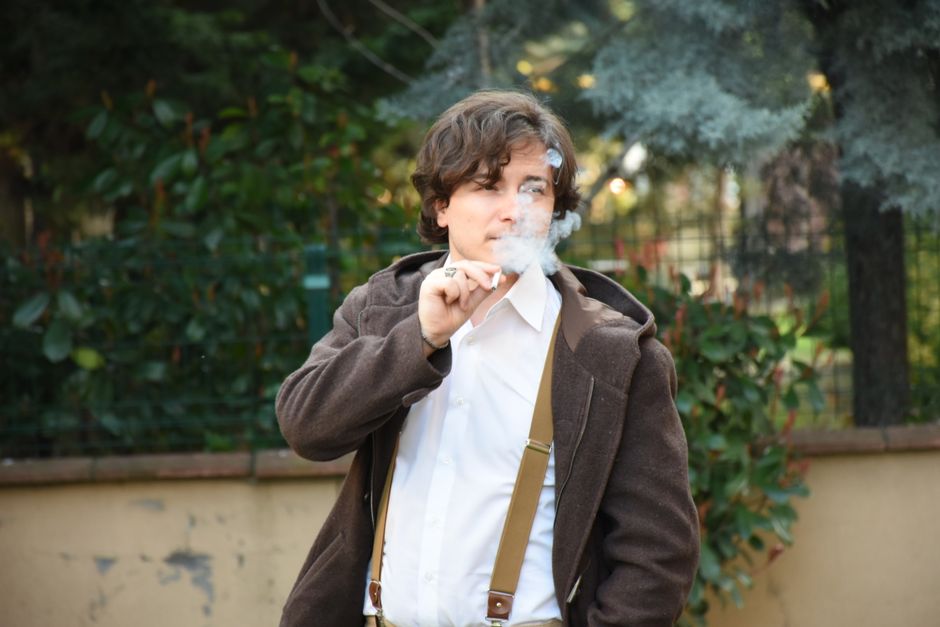 Hombre caucásico elegante con tirantes fumando en un entorno al aire libre frondoso.