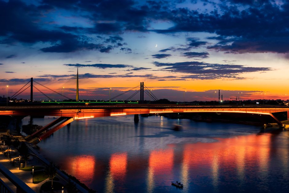 Vista escénica de un puente en Belgrado al atardecer con reflejos coloridos y una luna creciente.