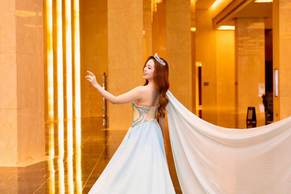 Una mujer con un vestido azul y una tiara posa en un pasillo lujoso y bien iluminado.